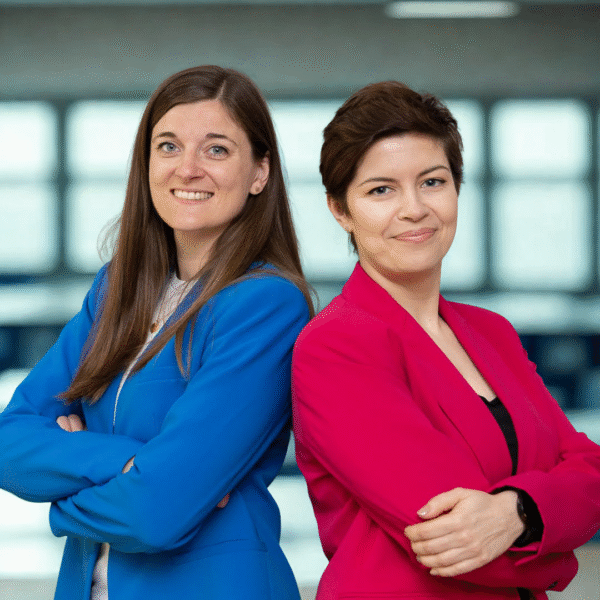 Daria i Kinga oparte o siebie plecami, uśmiechają się. Daria jest ubrana w marynarkę w kolorze chabrowym, a Kinga w jaskrawo różową.